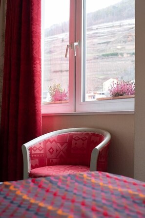 Habitació amb un llit, una finestra amb vista a un paisatge i una cadira amb coixí estampat.