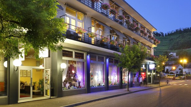 A multi-story hotel with balconies adorned with potted plants, large windows with curtains, and a well-lit facade. - The Originals City, Hotel de L'ange, Guebwiller (Guebwiller)