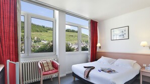 A hotel room with a large bed, a chair, a radiator, and a window with a view of a green landscape. - The Originals City, Hotel de L'ange, Guebwiller (Guebwiller)