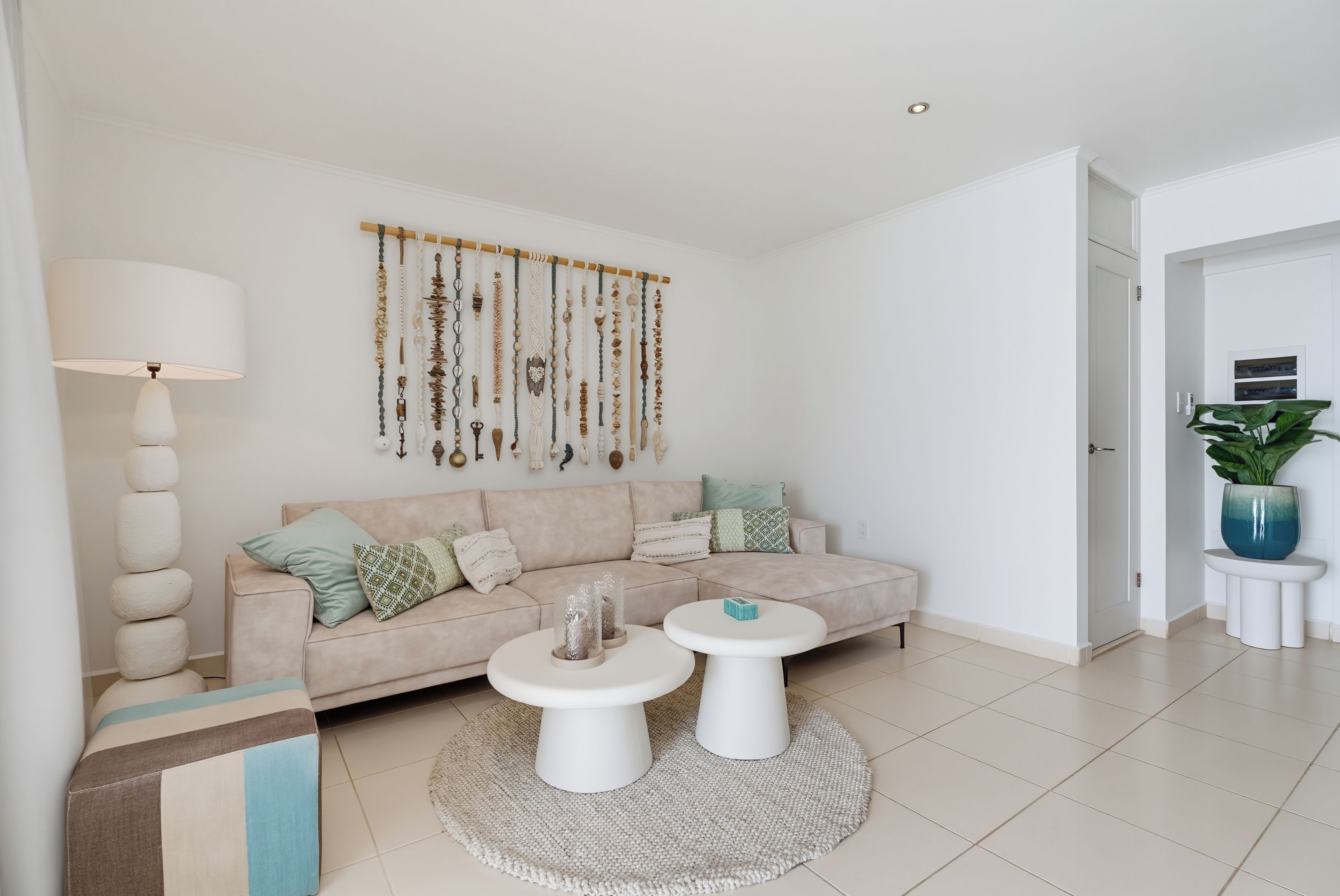 A modern living room with a beige sofa, two white coffee tables, a wall-mounted macrame wall hanging, and a potted plant.