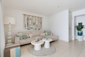 A modern living room with a beige sofa, two white coffee tables, a wall-mounted macrame wall hanging, and a potted plant.