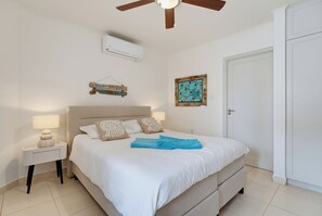 A bedroom with a bed, bedside tables, a ceiling fan, an air conditioning unit, and a door. - Oceanfront beach apartment Bellevue 2 (Kralendijk)