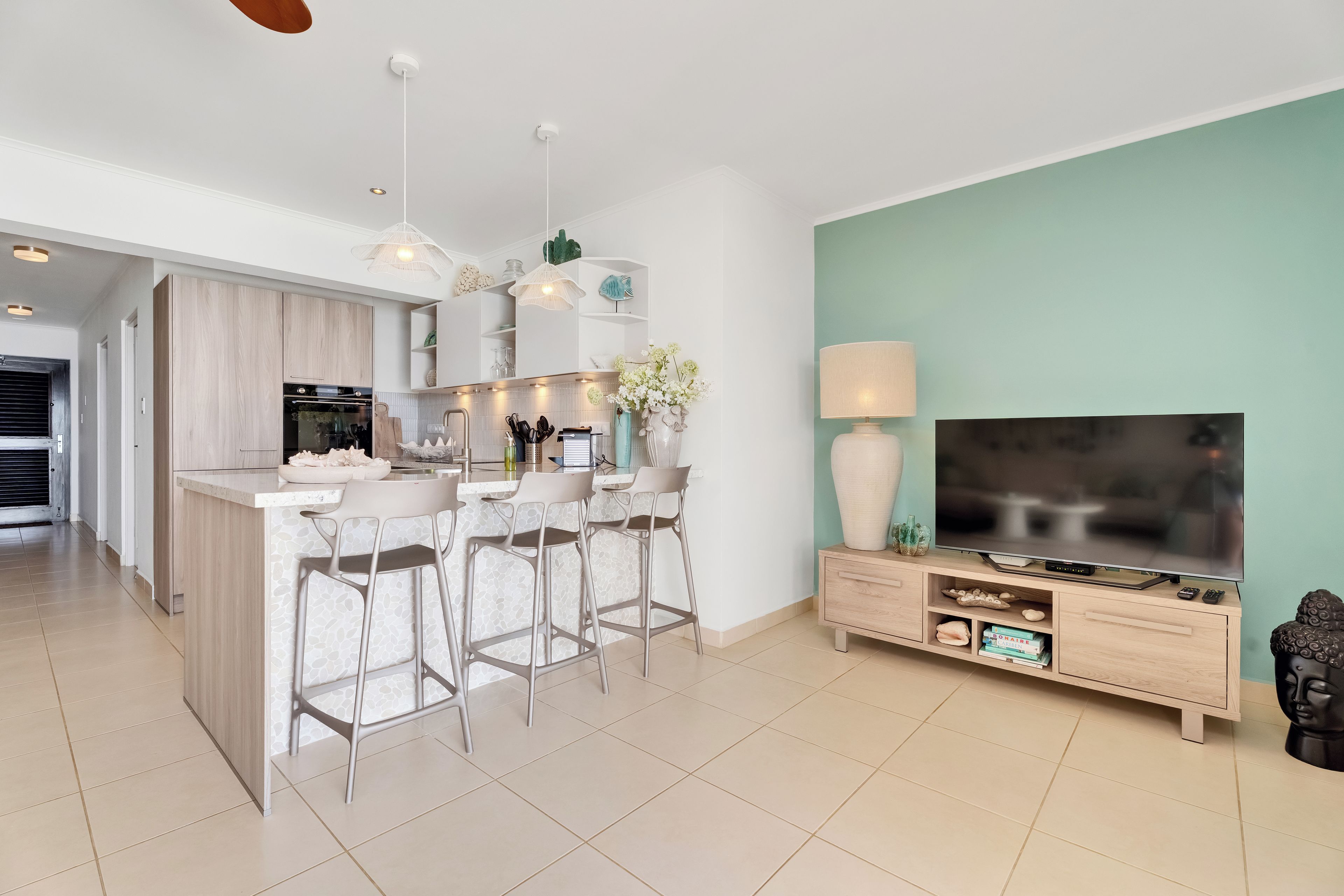 A modern kitchen with a central island, bar stools, a dining area, and a living room with a TV and entertainment unit.