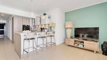 A modern kitchen with a central island, bar stools, a dining area, and a living room with a TV and entertainment unit.