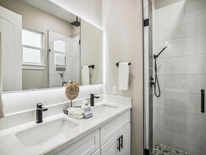 A modern bathroom with a large mirror, two sinks, a shower, and a towel rack. - Sandy Cheeks Surfside by Ryson Vacation Rentals (Freeport)