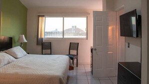 Ein Hotelzimmer mit einem Bett, zwei Stühlen, einem Fernseher und einem Fenster mit Blick auf Gebäude.