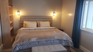 Ein Schlafzimmer mit einem hölzernen Kopfteil, zwei Nachttischen, einer Bank am Fußende des Bettes und einem Fenster mit Vorhängen.