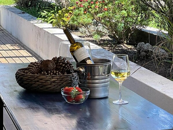 A bottle of wine, a glass of wine, a basket of pinecones, and a bowl of strawberries on an outdoor table.