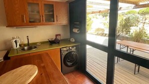 A compact kitchen with wooden cabinets, a sink, a washing machine, and a large window offering a view of the outdoors. - Omontinho - 4-bed House With Pool in Melides (Grandola)