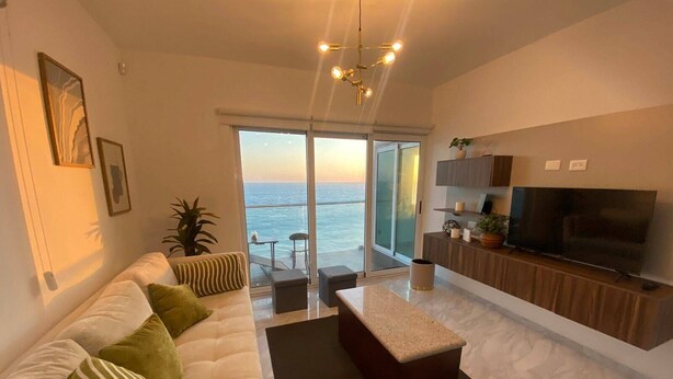 A modern living room with a sofa, coffee table, and TV. There is a balcony with a view of the sea. - Condos MDS7A ByTrvl2hm (Mazatlán)