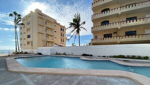 A swimming pool with a curved edge, surrounded by a concrete deck. There are palm trees and a white wall in the background. - Condos MDS7A ByTrvl2hm (Mazatlán)