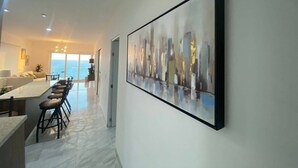A modern living room with a large abstract painting, a kitchen area with bar stools, and a view of the sea through large windows. - Condos MDS7A ByTrvl2hm (Mazatlán)