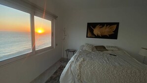 A bedroom with a large window overlooking the sea at sunset, a bed with a white comforter, a small bedside table, and a wall-mounted artwork. - Condos MDS7A ByTrvl2hm (Mazatlán)