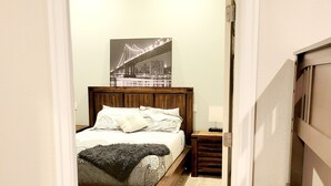 A bedroom with a bed, a nightstand, a lamp, and a cityscape painting on the wall. - Valley Views Apartment  (Happy Valley)