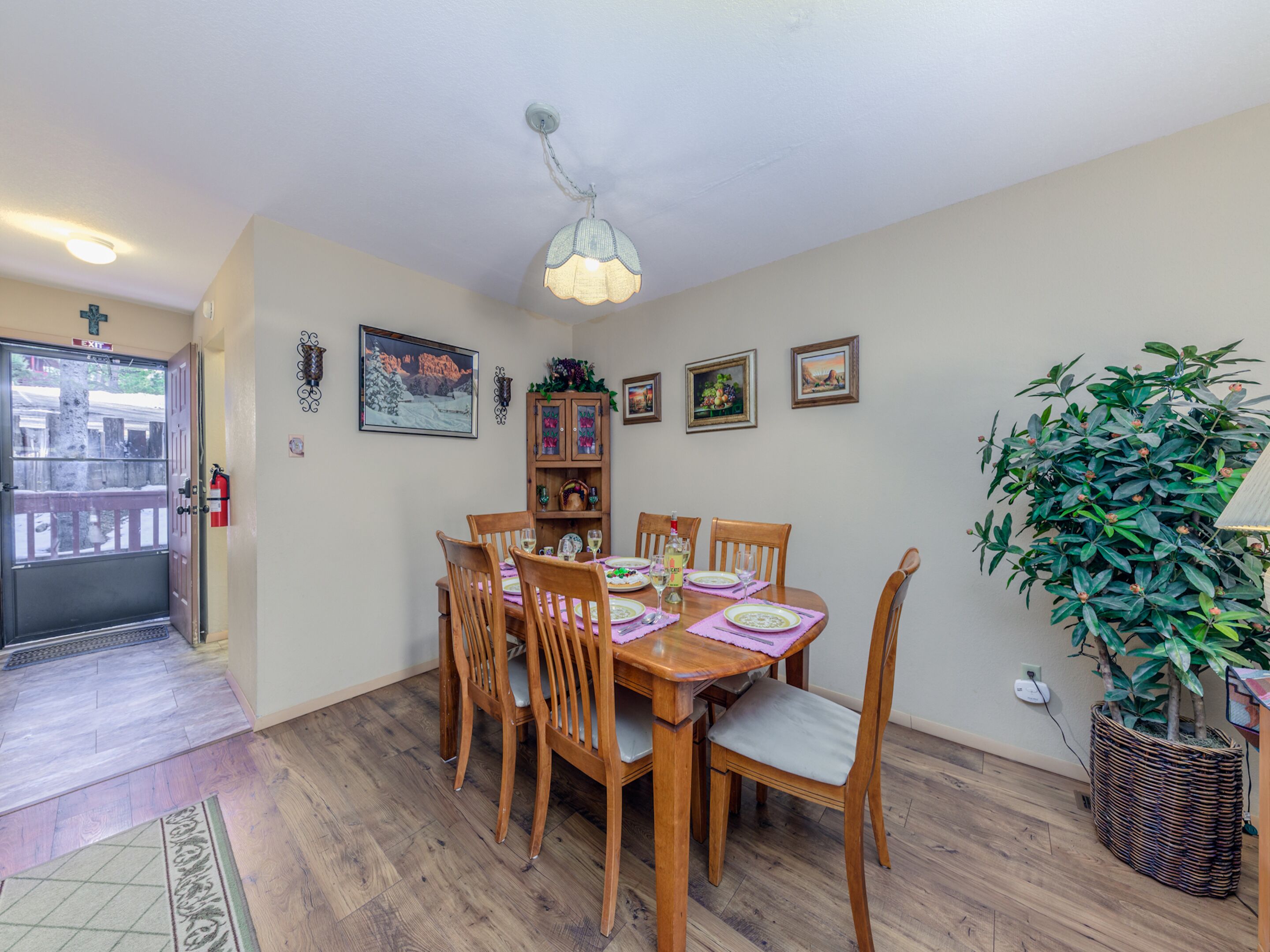 A dining area with a wooden table set for six, surrounded by chairs. There is a small cabinet with decorative items, a wall-mounted light fixture, and a potted plant to the side.