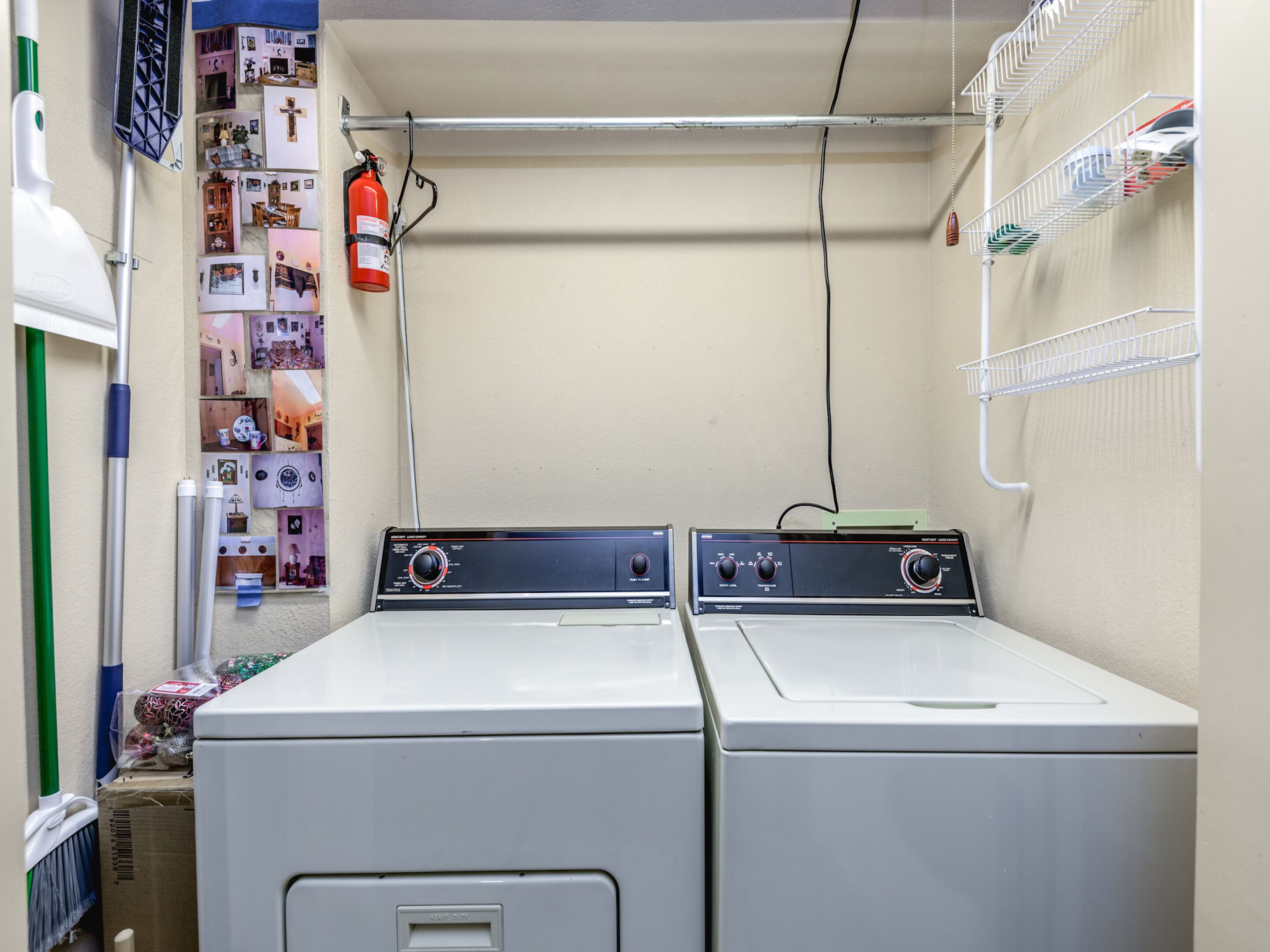 A laundry room with two front-loading washers and dryers, a fire extinguisher, and a wall-mounted shelf with various items.