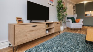 Ein modernes Wohnzimmer mit einem Flachbildfernseher auf einer Holzkonsole, einem gemusterten Teppich in Grün, einem grauen Sessel und einer Topfpflanze.