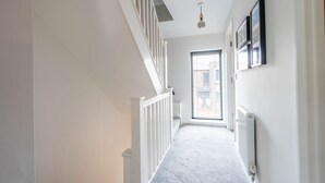Un couloir étroit avec un escalier blanc, un radiateur et une fenêtre équipée de stores.
