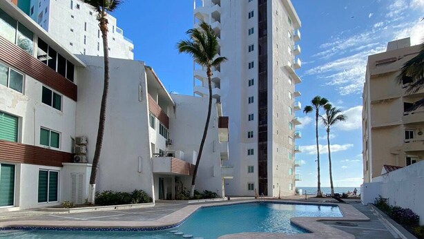 Área de piscina de resort com piscina azul cristalina, palmeiras e edifícios modernos com varandas.
