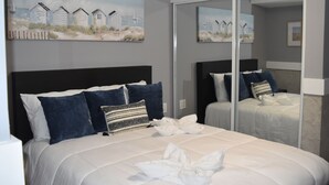 A bedroom with a large bed, two pillows, a white duvet, and a towel. There is a painting of beach huts on the wall and a mirror reflecting the room. - CALIZO VIP GUEST HOUSE  (Fajardo)