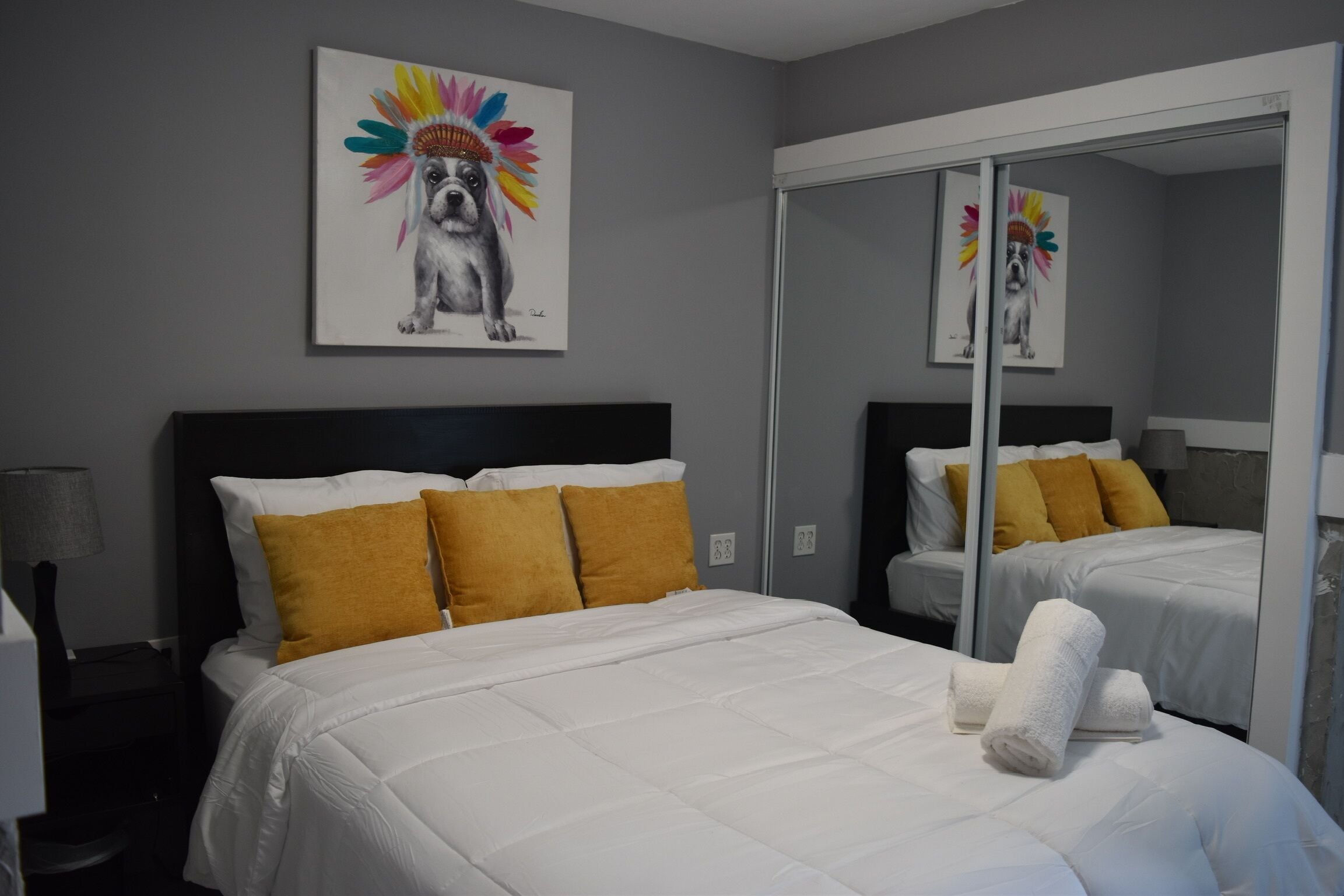 Habitación de hotel con cama, espejo y un cuadro de un perro usando un tocado colorido.