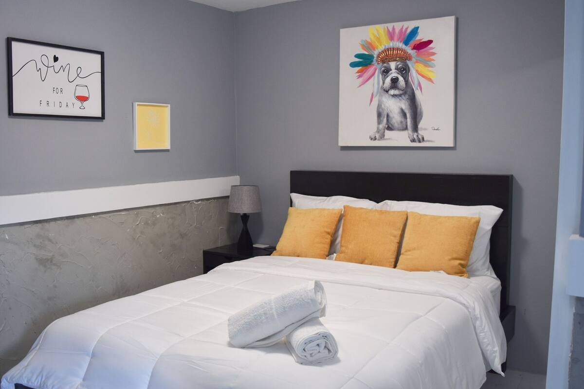 A neatly made bed with white bedding and a rolled towel, a nightstand with a lamp, and a framed picture of a dog with a colorful headdress on the wall.