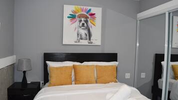 Une chambre d’hôtel avec un lit, une table de chevet avec une lampe et un tableau accroché au mur représentant un chien portant un couvre-chef coloré.