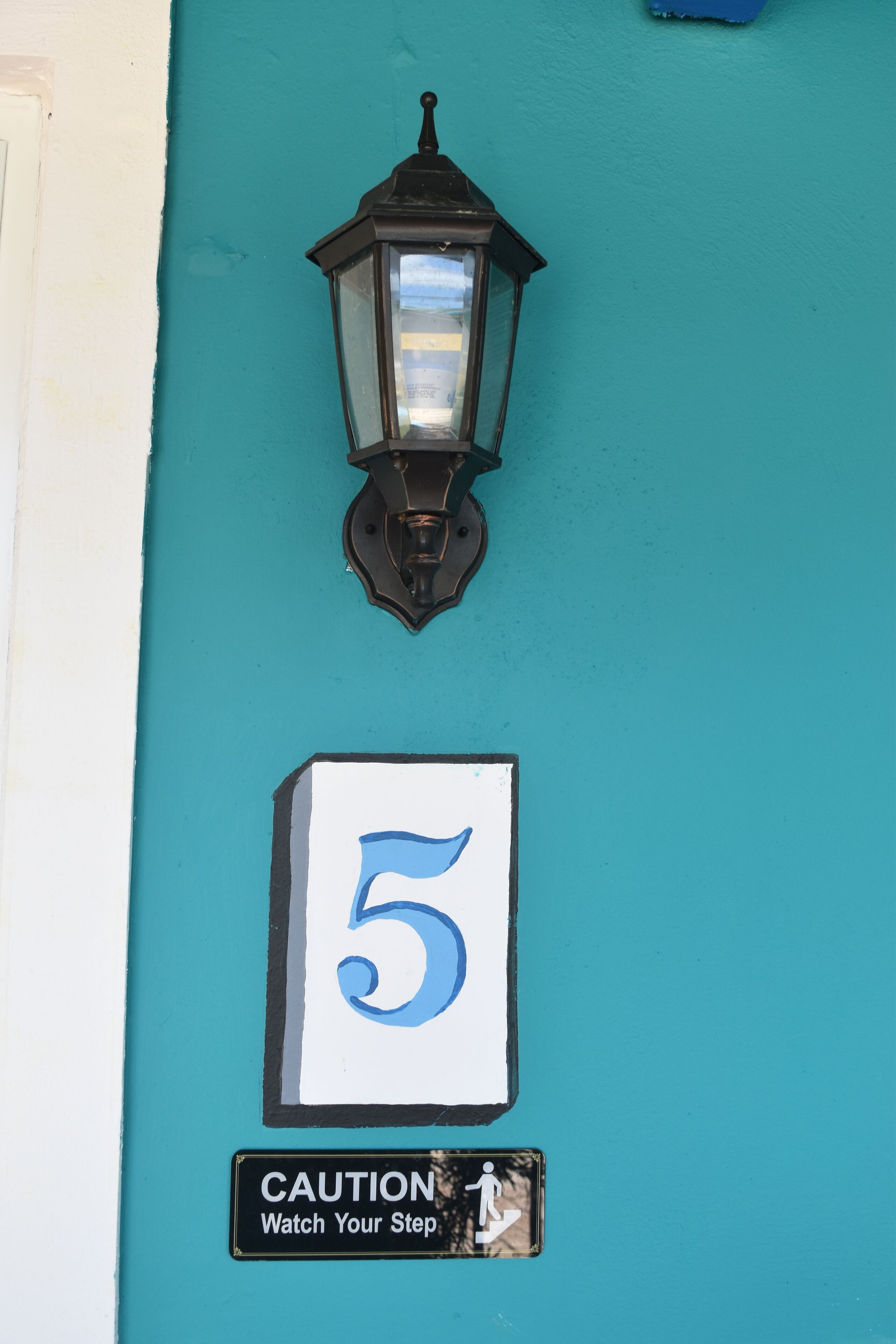 A teal wall with a black street lamp, a white sign with the number 5, and a caution sign with a pedestrian symbol.