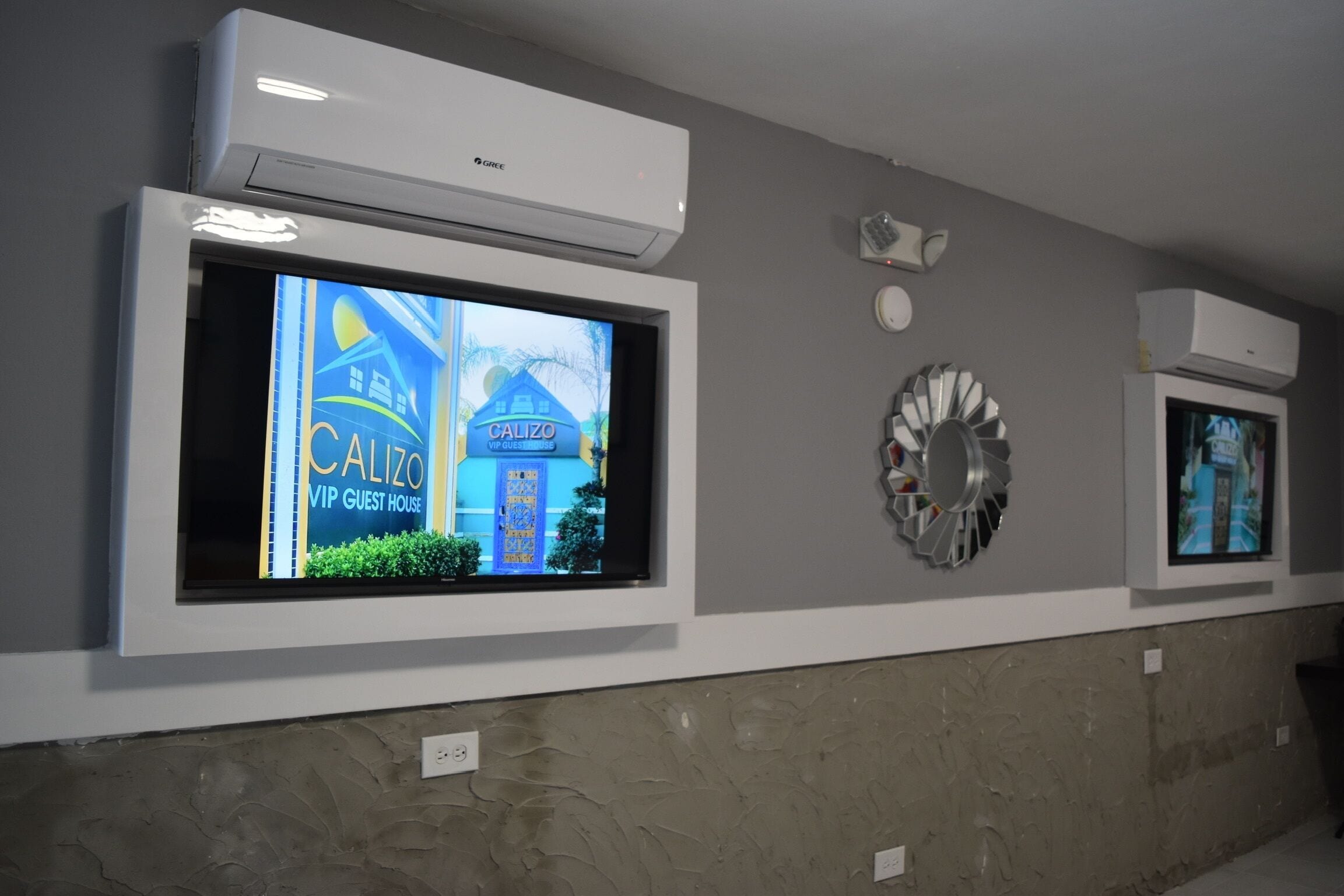 A hotel reception area with a large TV screen displaying a property advertisement, a decorative wall piece, and an air conditioning unit.