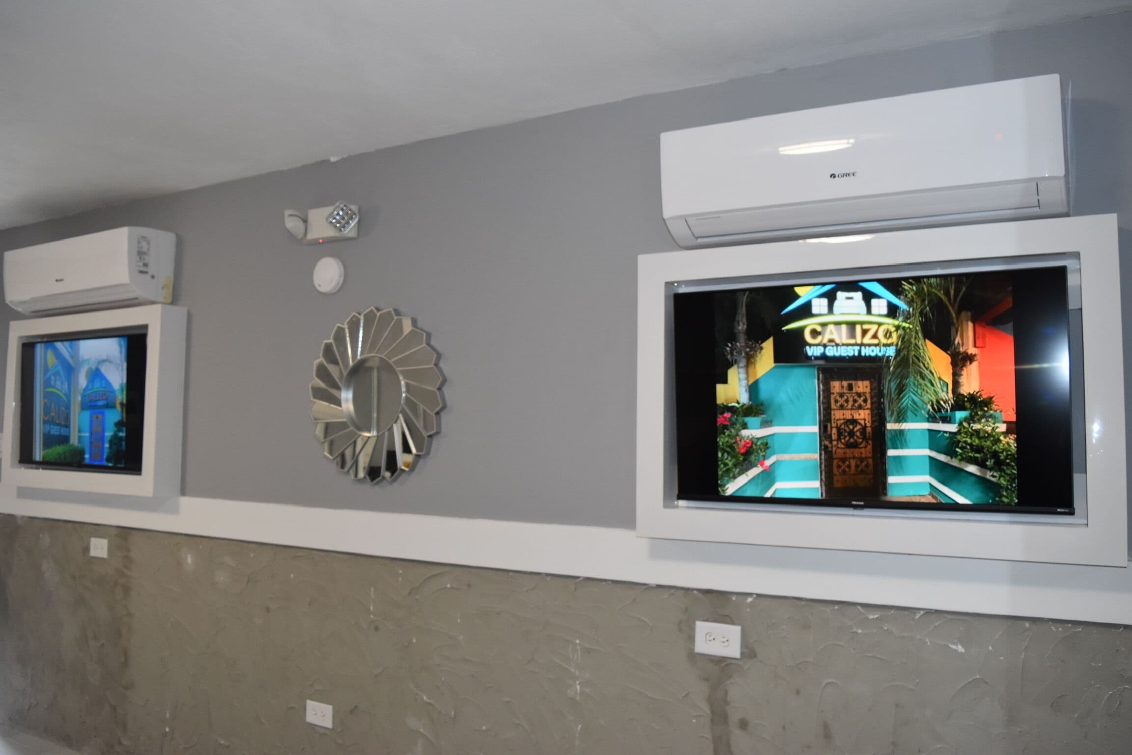 Pared de hotel con un televisor que muestra un letrero 'Calizo', un equipo de aire acondicionado y un adorno de metal.