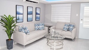 A modern living room with a white sofa, two patterned pillows, a glass coffee table, and a potted plant. - CALIZO VIP GUEST HOUSE  (Fajardo)