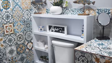 Un baño con un inodoro blanco, una estantería con un cartel que dice 'love is us', un florero con flores blancas y una decoración de pez.