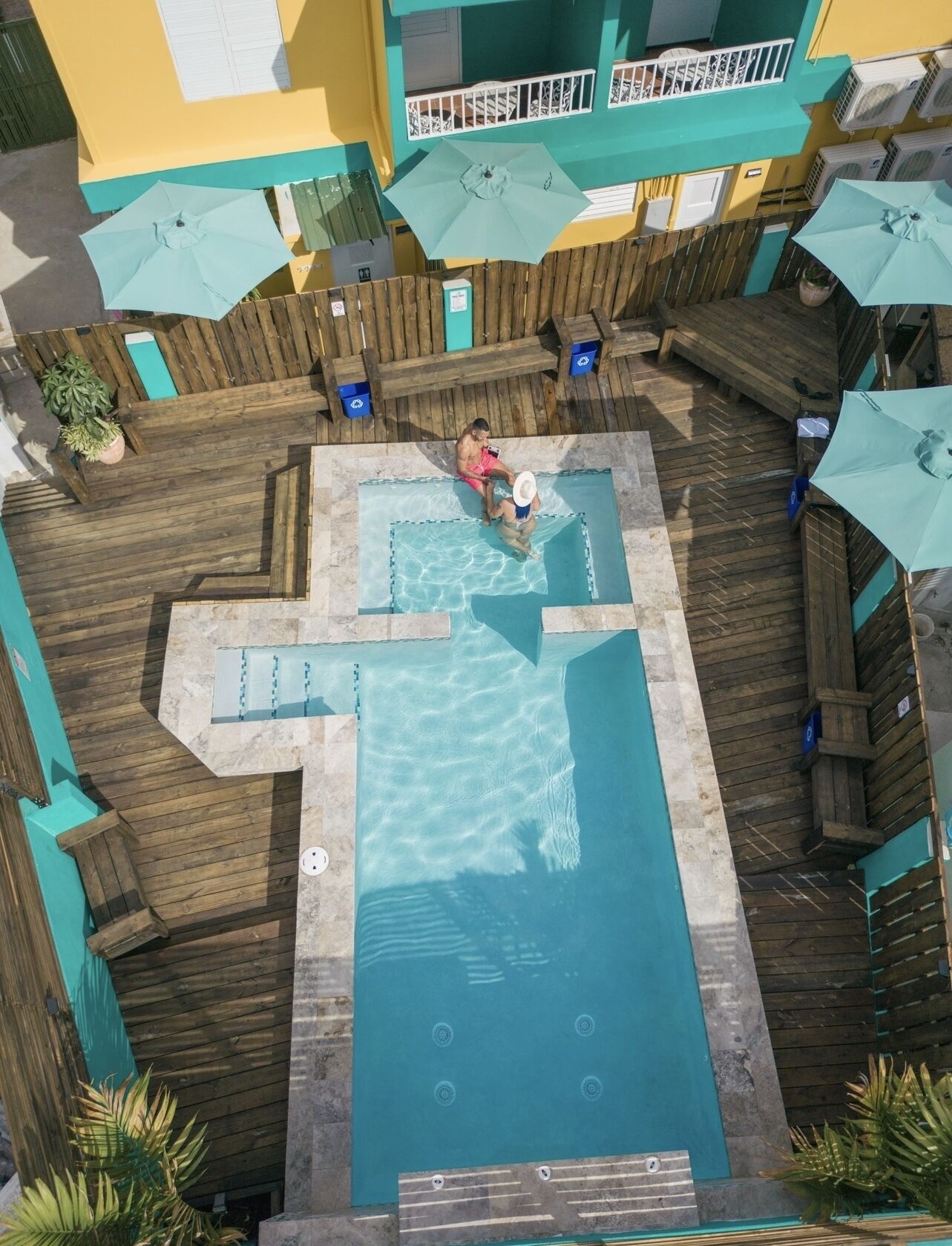 A rooftop pool with a wooden deck, surrounded by potted plants and umbrellas.