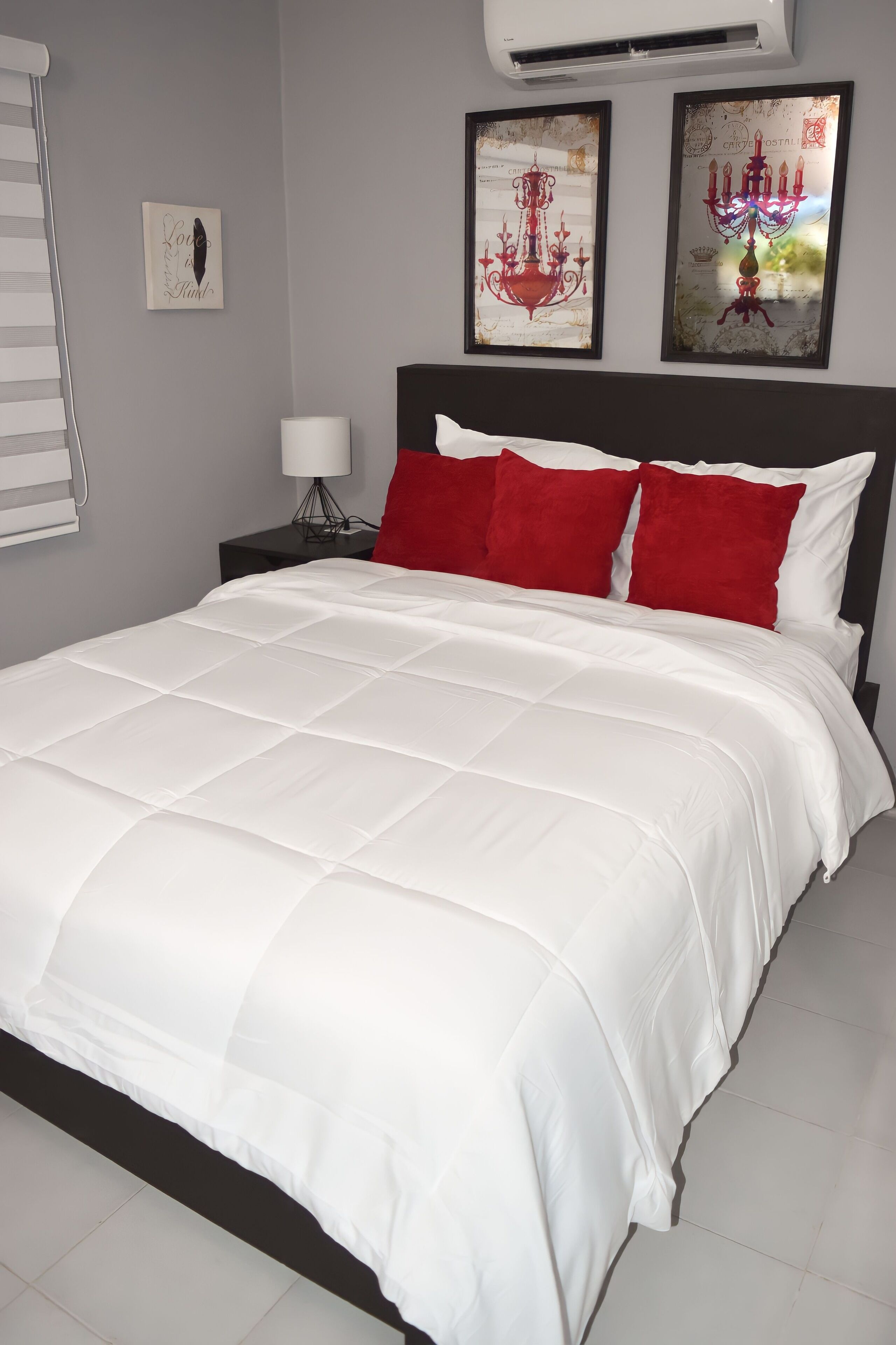 A neatly made bed with white bedding and red pillows, a bedside lamp, and two wall art pieces above it.