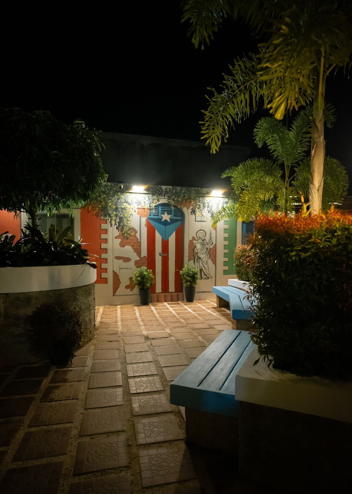 An outdoor patio area with tiled flooring, benches, potted plants, and a mural on the wall.