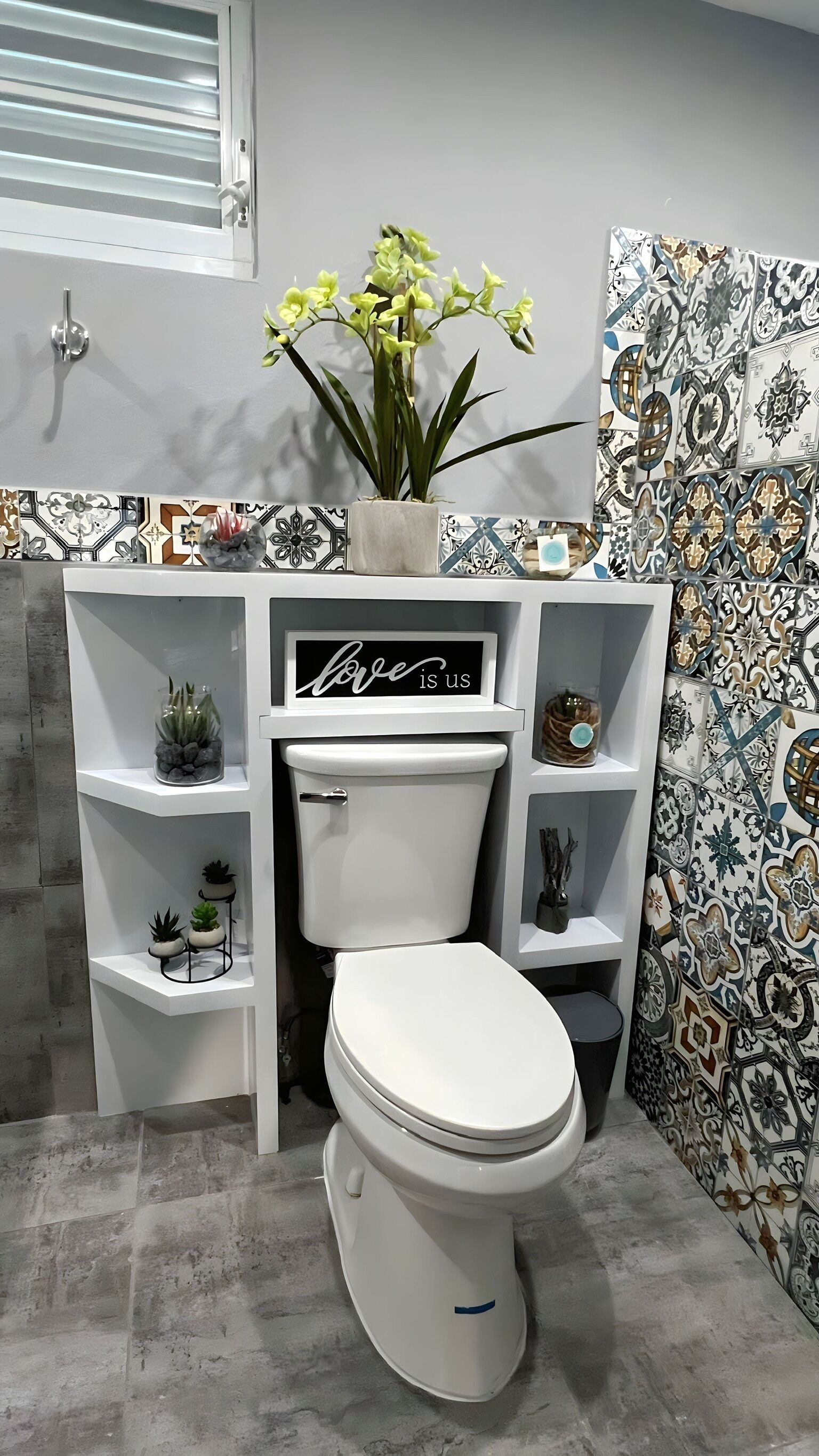 Un baño moderno con inodoro, estantes con plantas, un letrero decorativo y baldosas estampadas.