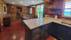 A kitchen with wooden cabinets, a marble countertop, and a dining area with a table and chairs. - Knotty & Nice Cottage Mountain Views, Near Tail of the Dragon, & Fontana Village (Fontana Dam)