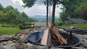 A fire pit with logs, surrounded by rocks and gravel, in a natural outdoor setting with trees and a distant house. - Knotty & Nice Cottage Mountain Views, Near Tail of the Dragon, & Fontana Village (Fontana Dam)