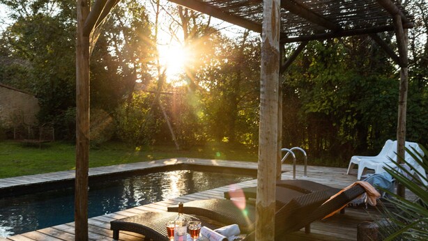 Una terraza de madera con piscina, sillas de descanso y una mesa con bebidas y un periódico.