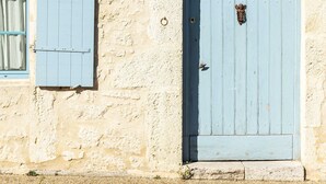 Un edificio con fachada de piedra clara, una puerta azul con aldaba y una ventana con contraventanas azules.
