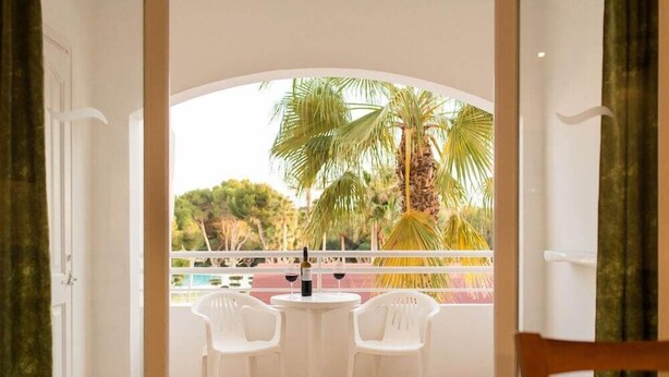 A balcony with two white plastic chairs and a table, overlooking a pool and palm trees. - Apartamentos Vista Playa I (Ciutadella de Menorca)