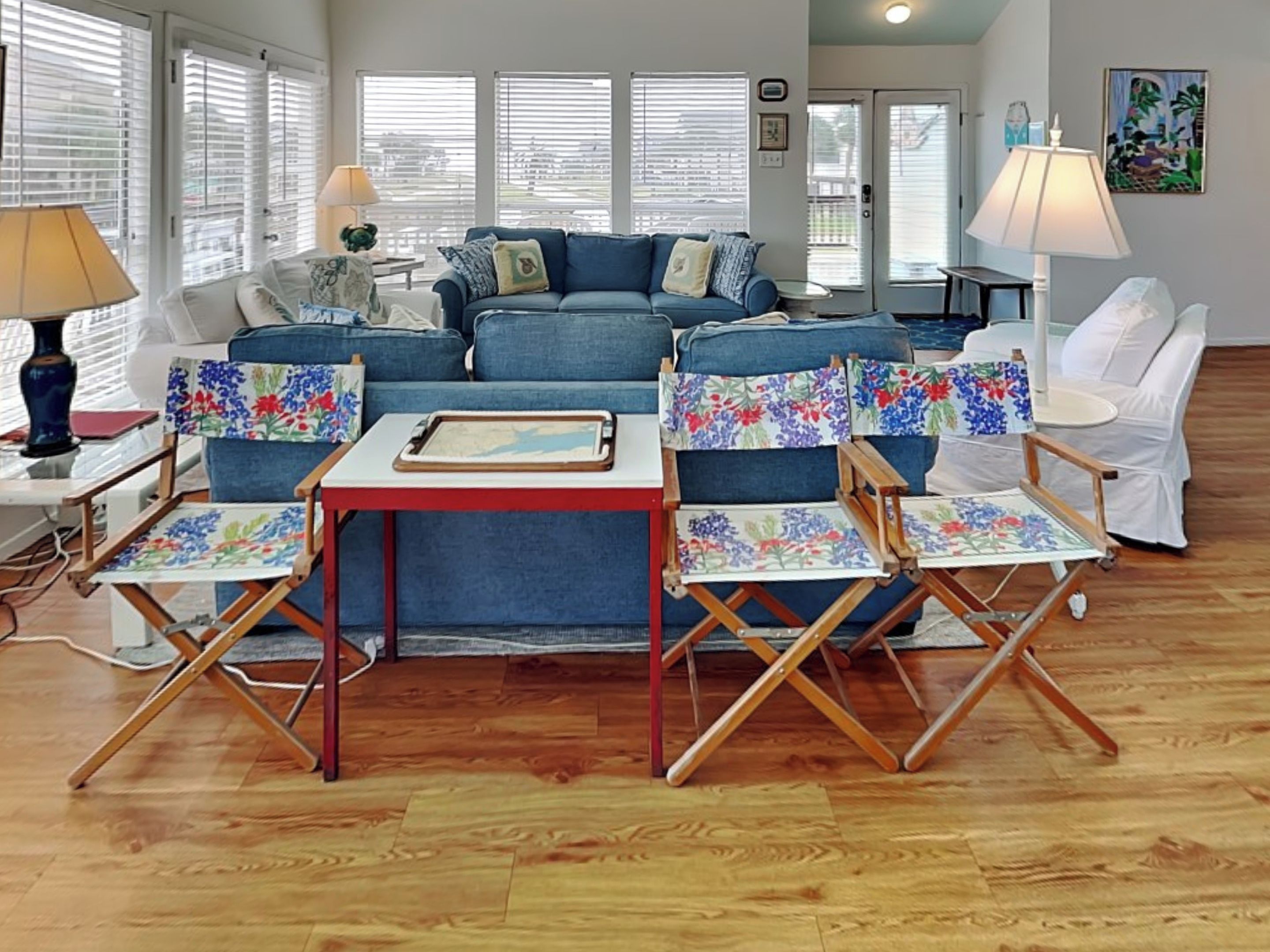 Uma sala de estar com um sofá azul, poltronas florais, uma mesa pequena com um tabuleiro e uma cadeira branca.