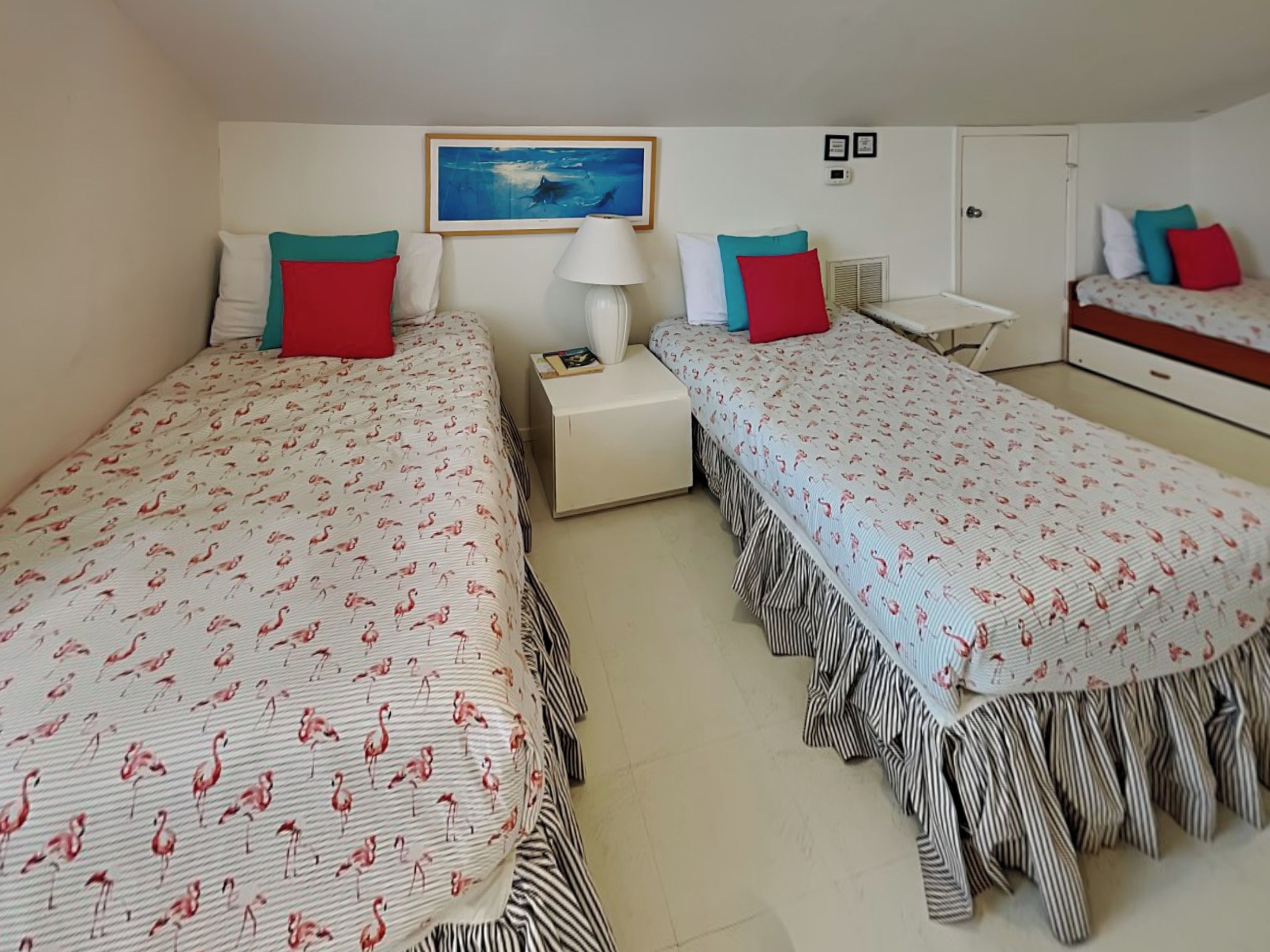 Een hotelkamer met twee bedden, elk met een gestreept dekbed en rode kussens. Er staat een nachtkastje met een lamp, een kleine tafel en een lijst met een afbeelding aan de muur.