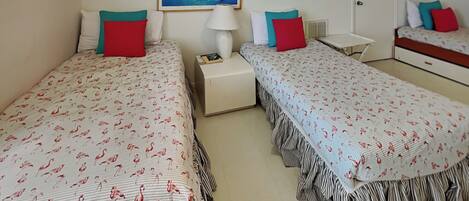 A hotel room with two beds, each with a striped bedspread and red pillows. There is a nightstand with a lamp, a small table, and a framed picture on the wall.