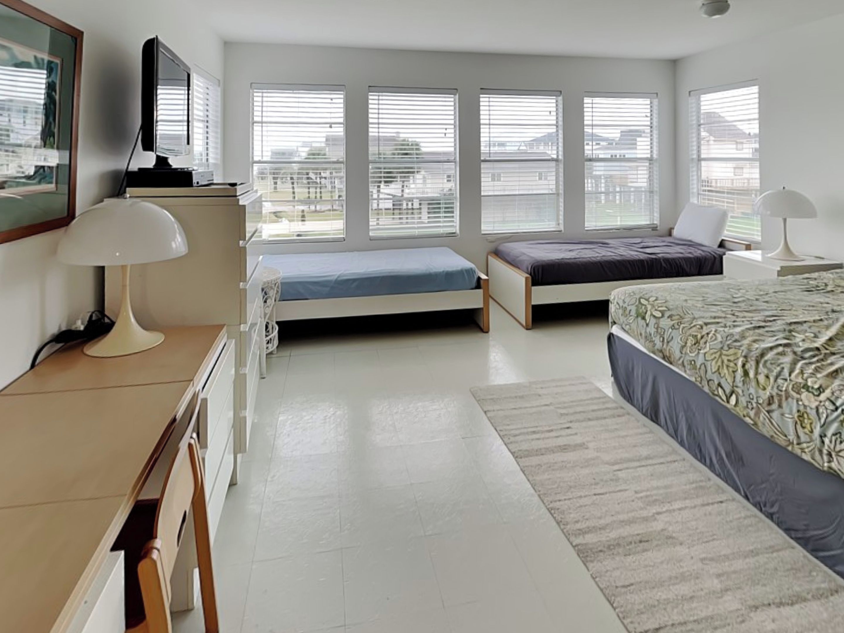 A bedroom with two single beds, a desk with a lamp, a TV, and large windows with blinds.