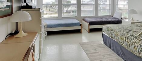 A bedroom with two single beds, a desk with a lamp, a TV, and large windows with blinds.