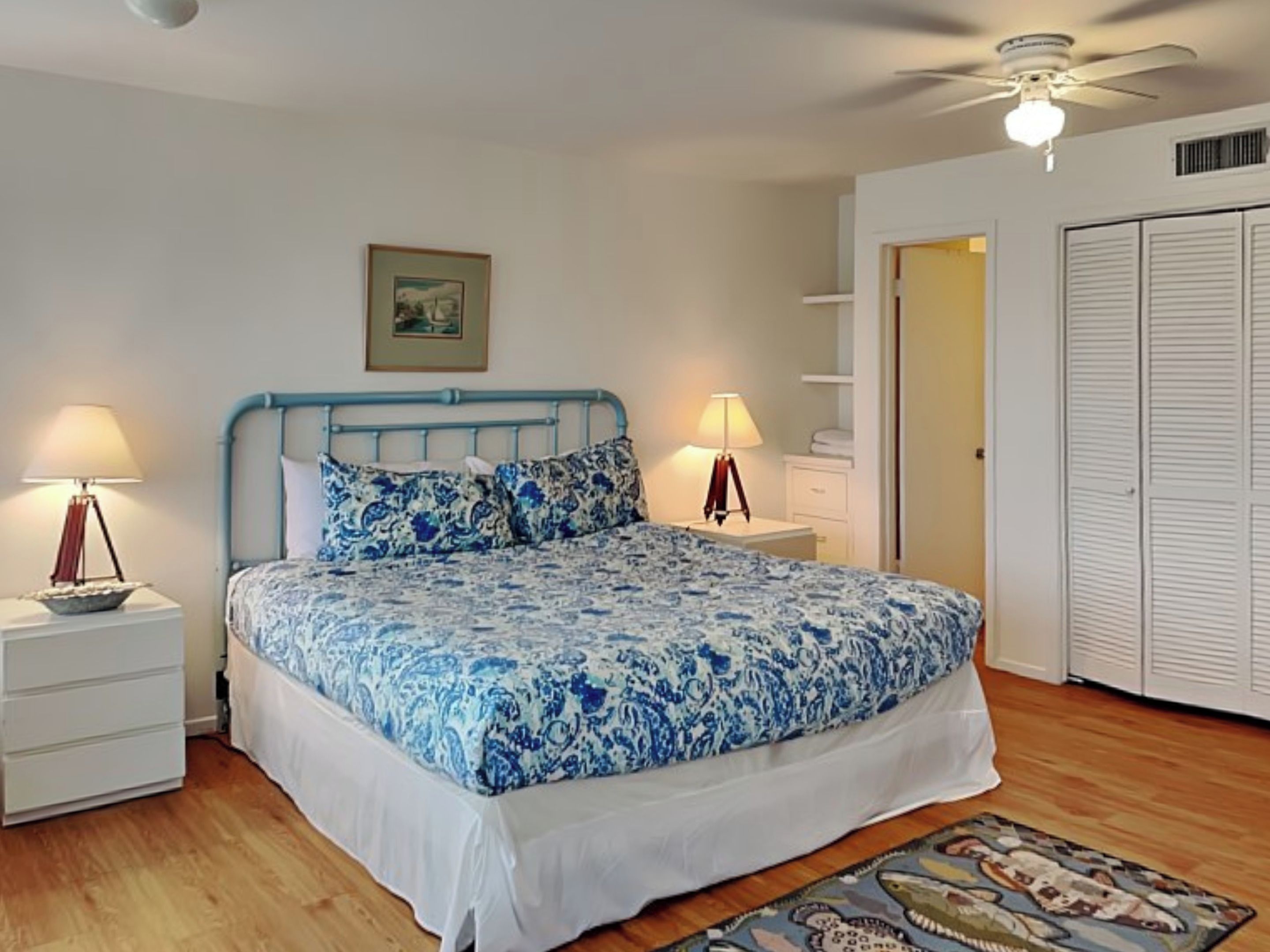 A bedroom with a bed, two bedside tables, a ceiling fan, a wardrobe, and a door.