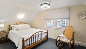 A bedroom with a wooden bed, a chair, a bedside table, a window with blinds, and a decorative wall piece. - Patty's Place - Private and Convenient Apartment near Downtown (Frederick)