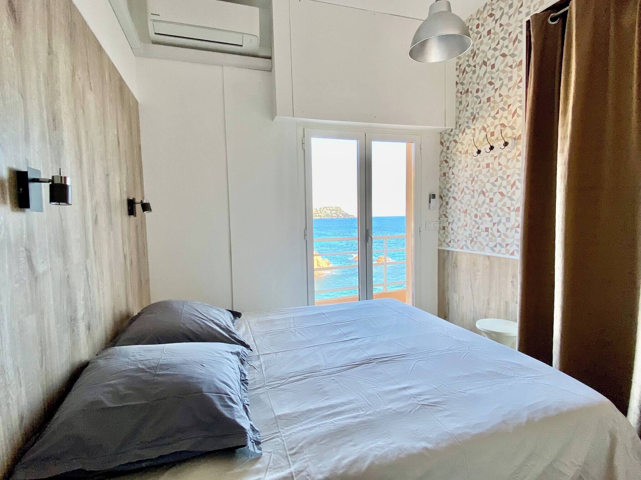 A bedroom with a bed, a window with a view of the sea, a ceiling fan, and a wall-mounted light fixture.