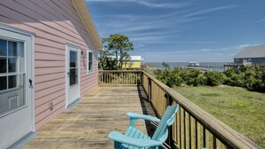 Eine Holzterrasse mit zwei hellblauen Adirondack-Stühlen, ein rosafarbenes Gebäude mit weißer Verzierung und im Hintergrund Meer und eine Pier.